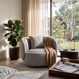 A modern round lounge chair in light gray bouclé fabric with a matching pillow and a tan throw blanket, styled in a bright, sunlit living room with a large window, potted plant, and side table.